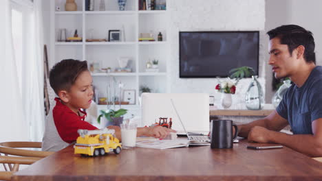 Hispanic-father-and-son-working-opposite-at-dining-room-table,-side-view,-panning,-left-to-right