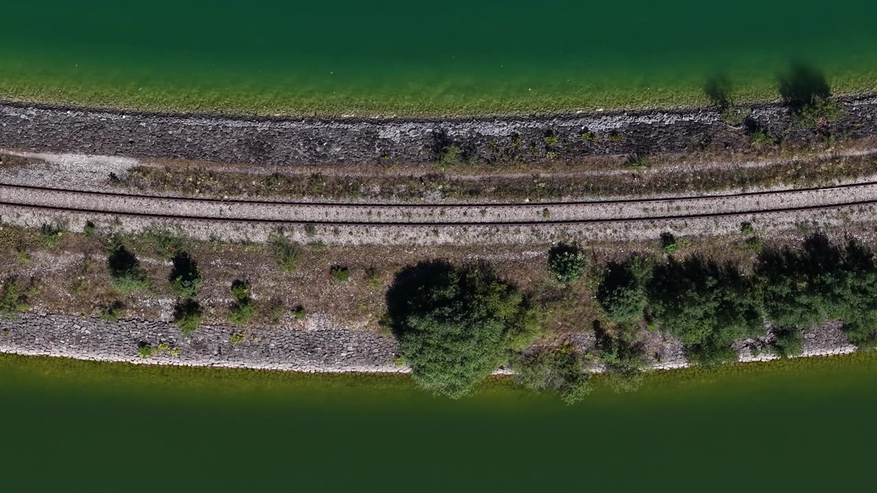 An aerial view shows parallel train tracks running alongside a rocky embankment and serene green water.