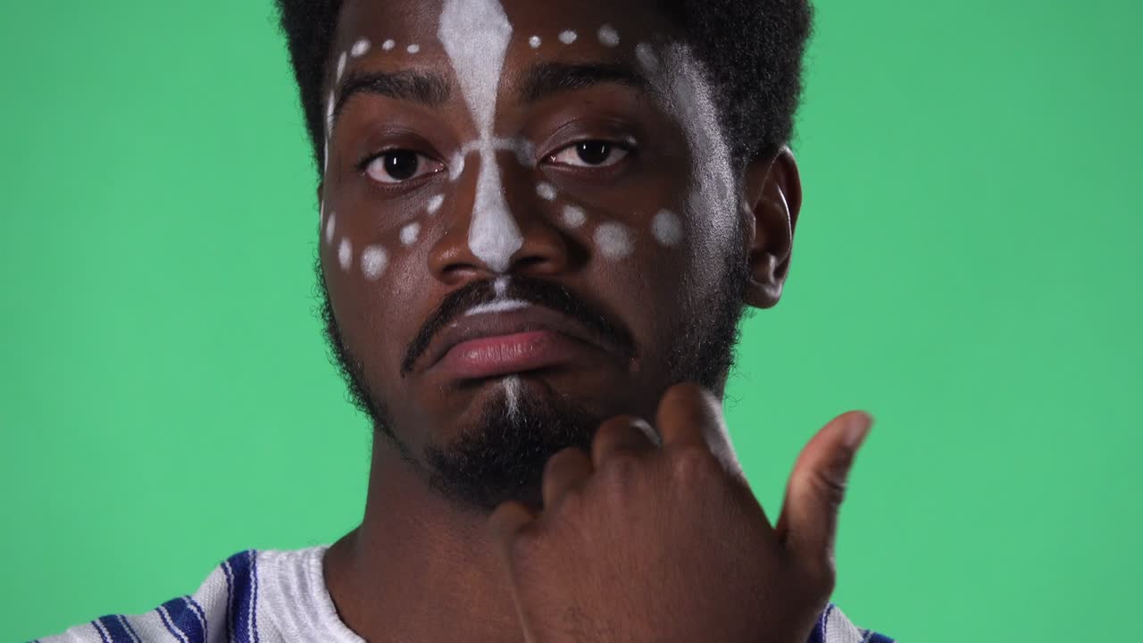 젊은 아프리카계 미국인 남성이 카메라를 바라보며 정보를 듣고, 얼굴에 흑인 백인 패턴을 가진 남성이 스튜디오에서 녹색 스크린에 포즈를 취하고 있습니다.