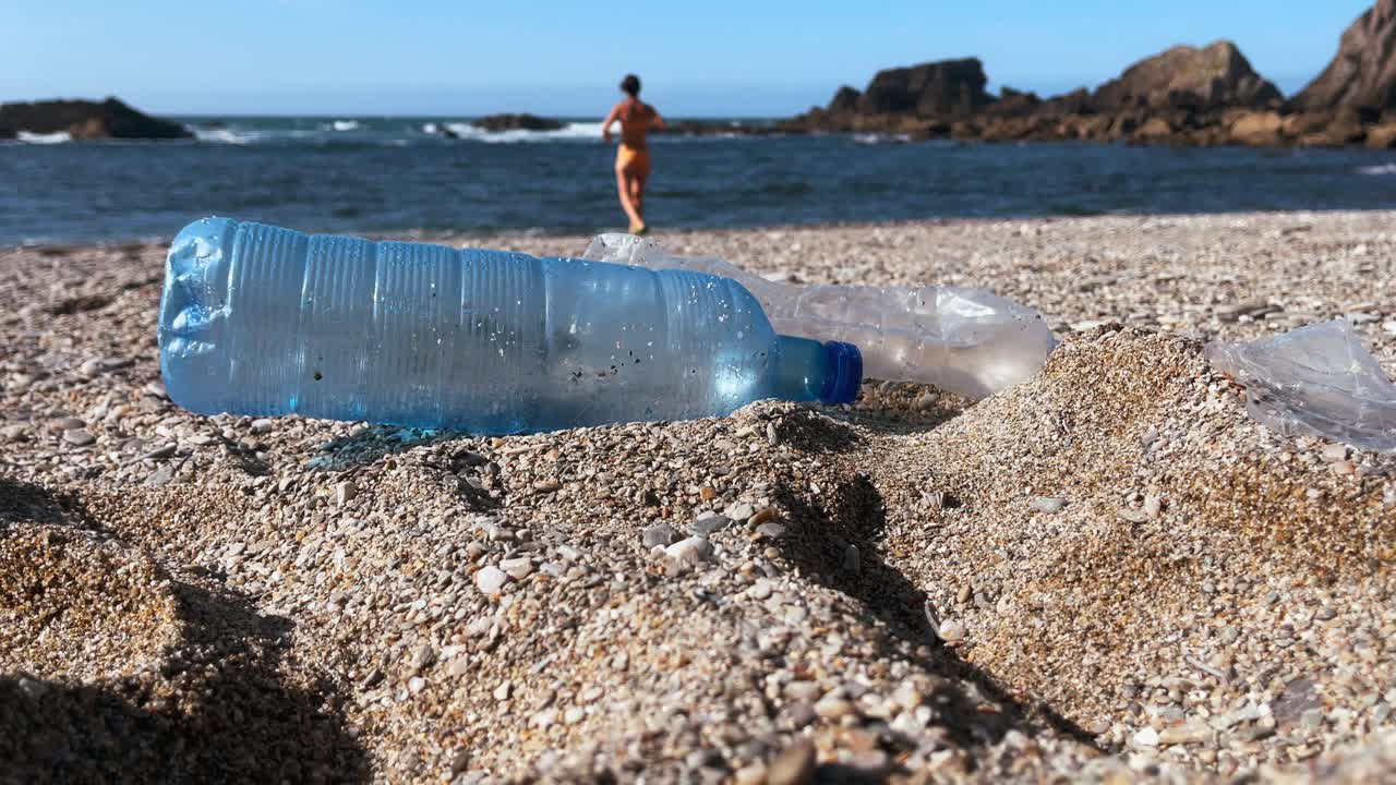 botellas de plástico ensucian una playa de arena con una persona corriendo hacia el océano