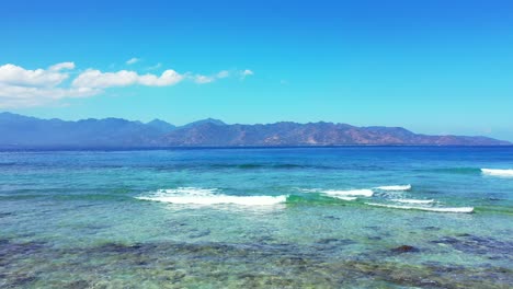 beautiful seashore with shallow calm water over coral reefs and rocky seabed where white waves splashing and foaming with blue sea and mountains background