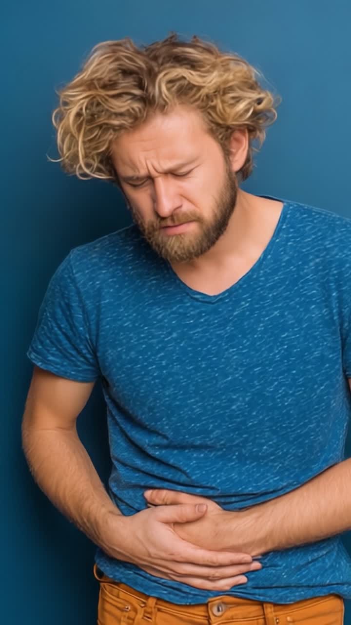 Un uomo che prova disagio e dolore, afferrandosi lo stomaco mentre trasmette un senso di angoscia su uno sfondo blu solido.