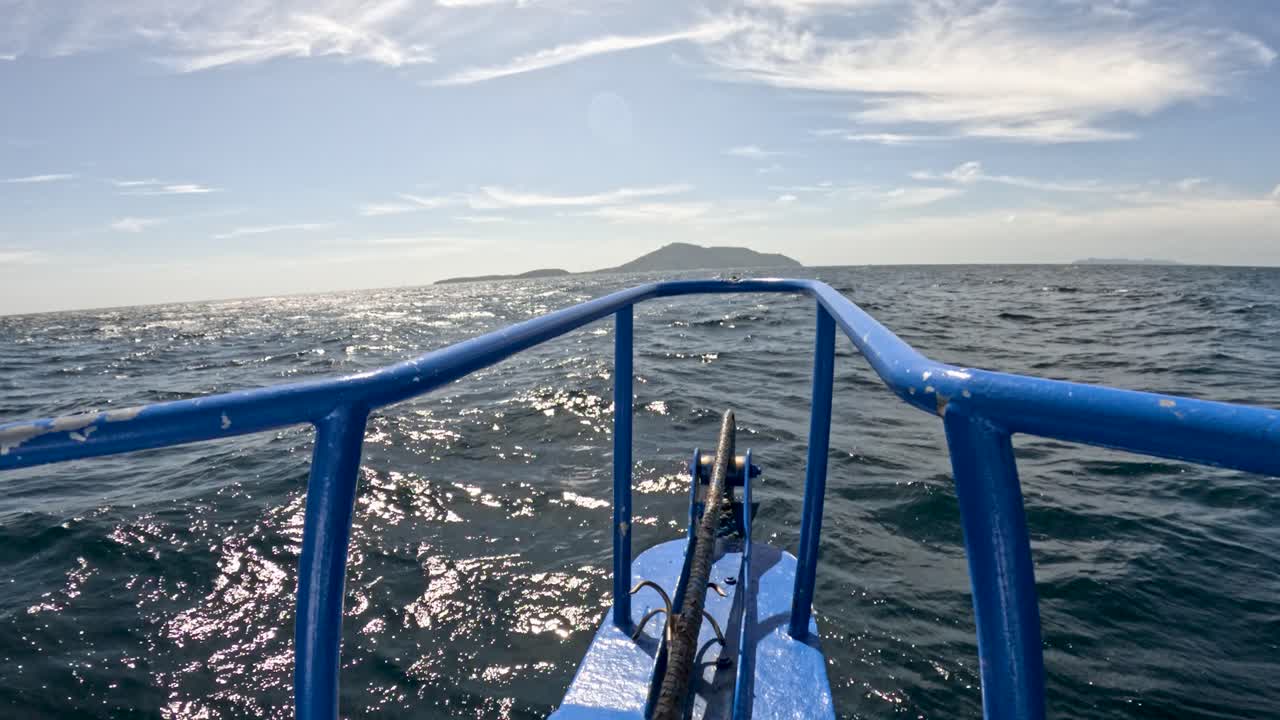 Blue boat moves through sparkling sea toward distant island, midday sunlight, steady forward camera view