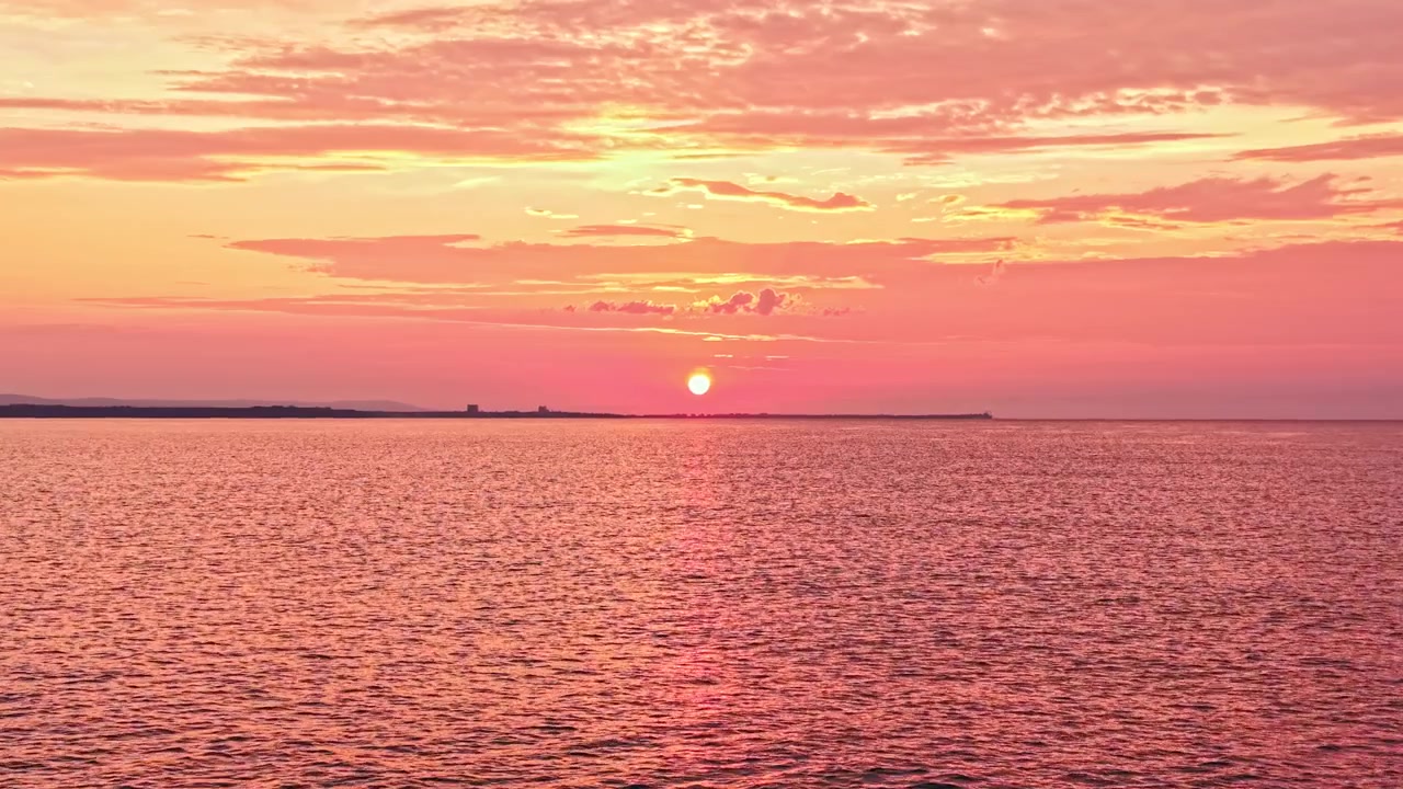 Solnedgang over vandet med lyserøde og orange farver, der fylder himlen og reflekteres på den rolige overflade, set fra kysten i aftentimerne.