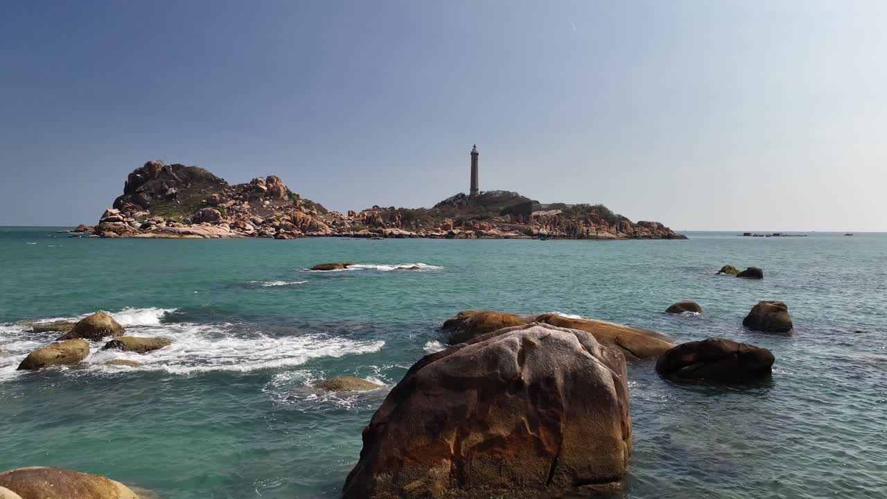 Rocky island with tall lighthouse stands in calm blue sea, waves gently crashing, natural coastline scenery under clear sky daytime in Vietnam coastal landscape