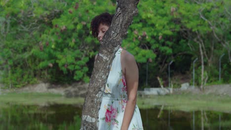 Bonita-Chica-De-Cabello-Rizado-Rojo-Caminando-En-Un-Parque-Usando-Un-Vestido-Largo-De-Flores-Con-Un-Estanque-En-El-Fondo