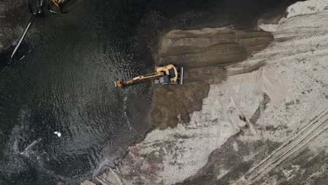 drone gets closer to a yellow excavator as it cleans up a river and piles dirt on shore