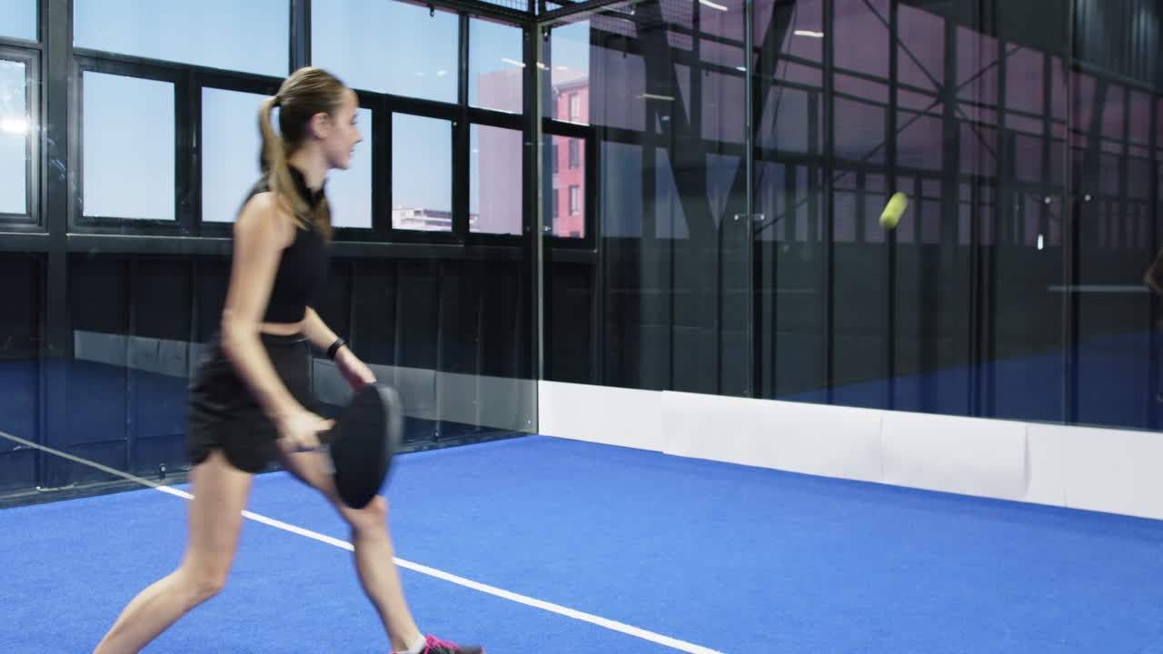 Young woman playing padel tennis on indoor court, focusing on hitting ball
