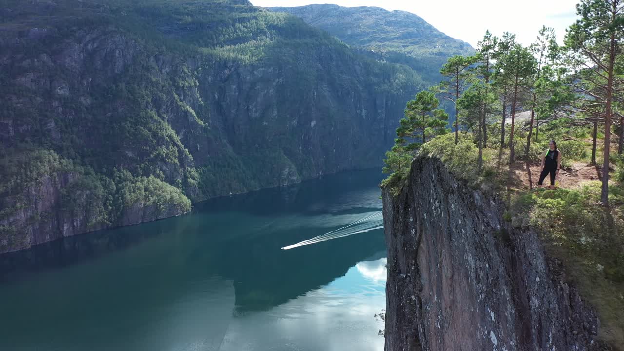 Woman enjoying spectacular view from edge of tall mountain cliff Slottet in Modalen Norway - Forward moving aerial close to cliff in stunning fjord landscape with boat passing on fjord below - Norway