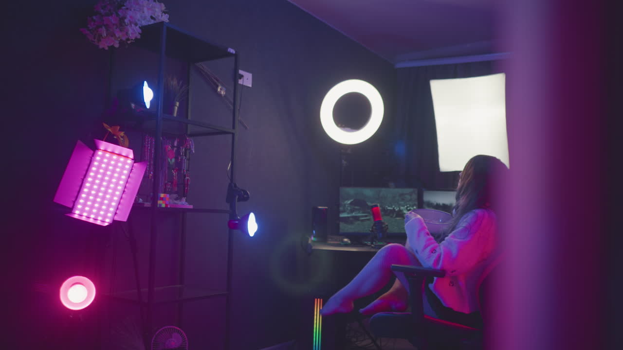 Ergonomic gaming chair positioned before illuminated monitor and ring light, empty amid purple and pink neon studio setting, inviting seating for upcoming gaming as woman approaches to sit down