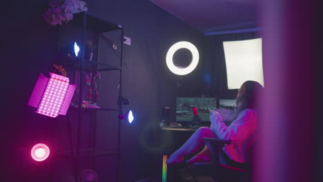 ergonomic gaming chair positioned before illuminated monitor and ring light, empty amid purple and pink neon studio setting, inviting seating for upcoming gaming as woman approaches to sit down