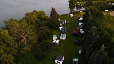 a row of portable ice fishing shanty cabin huts beached on grassland near a lake in the summer time