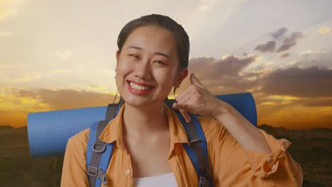 close up of asian female hiker with mountaineering backpack smiling and showing call me gesture while standing on the top of mountain during sunset time