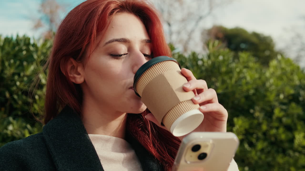 Young Red-Haired Woman Drinking Coffee and Using Smartphone Outdoors