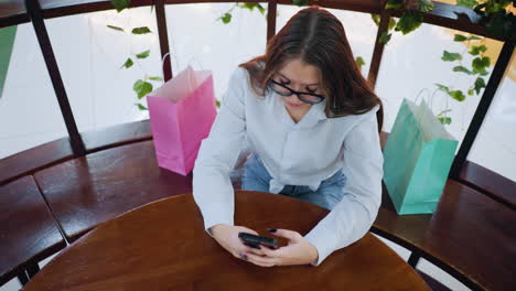 overhead view of woman chatting with friend over phone, hand placed on round table with shopping bags beside, in a vibrant, well-lit cafe setting