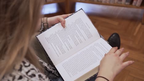 Young-blonde-girl-reading-a-book,-sitting-on-a-chair-in-library,-high-angle-closeup