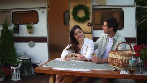 loving european couple man and woman drinking white wine while sitting together at wooden table outdoors. man caressing hair of his beloved. stylish, static wheels house on background