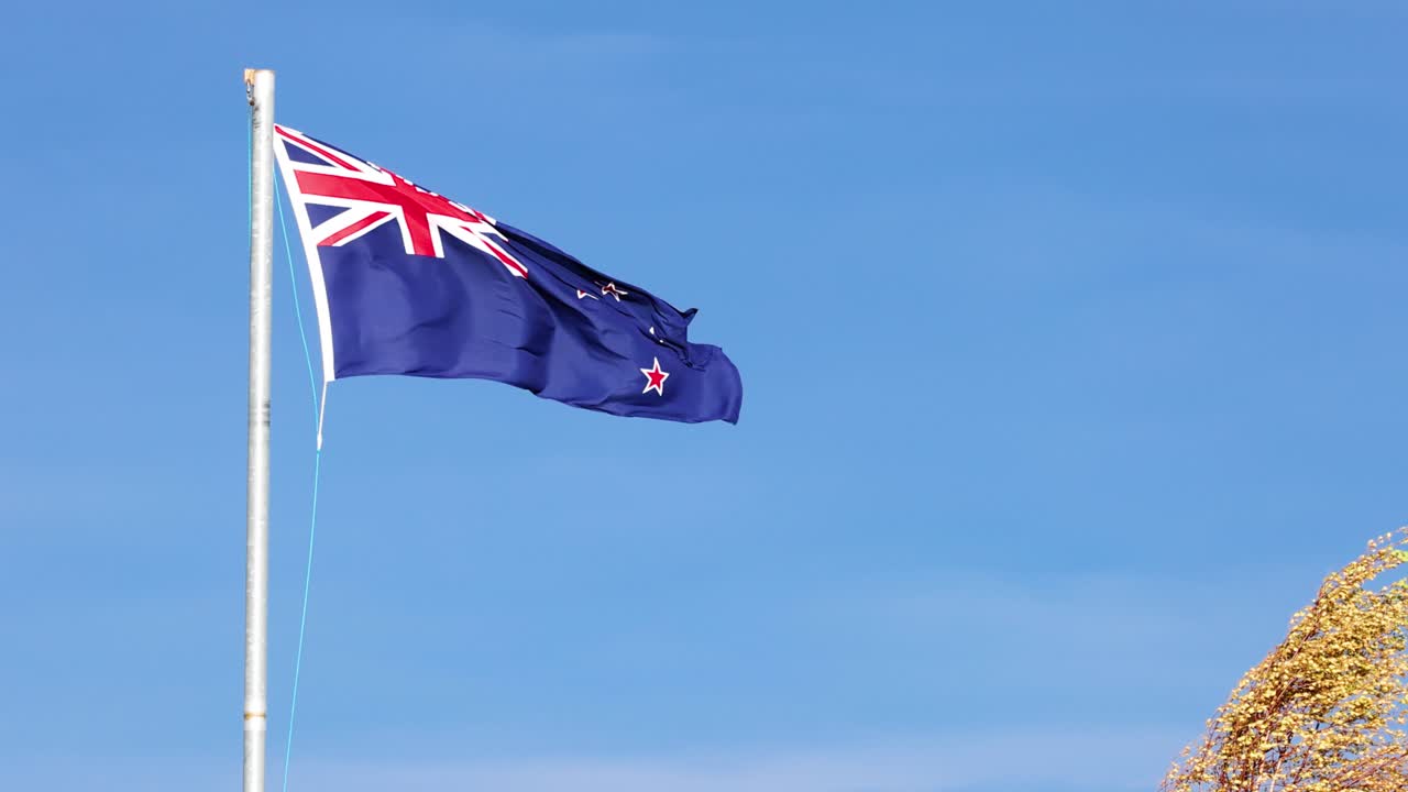A New Zealand flag flutters vigorously on a tall flagpole against a clear blue sky, with rocky terrain visible, captured in bright daylight