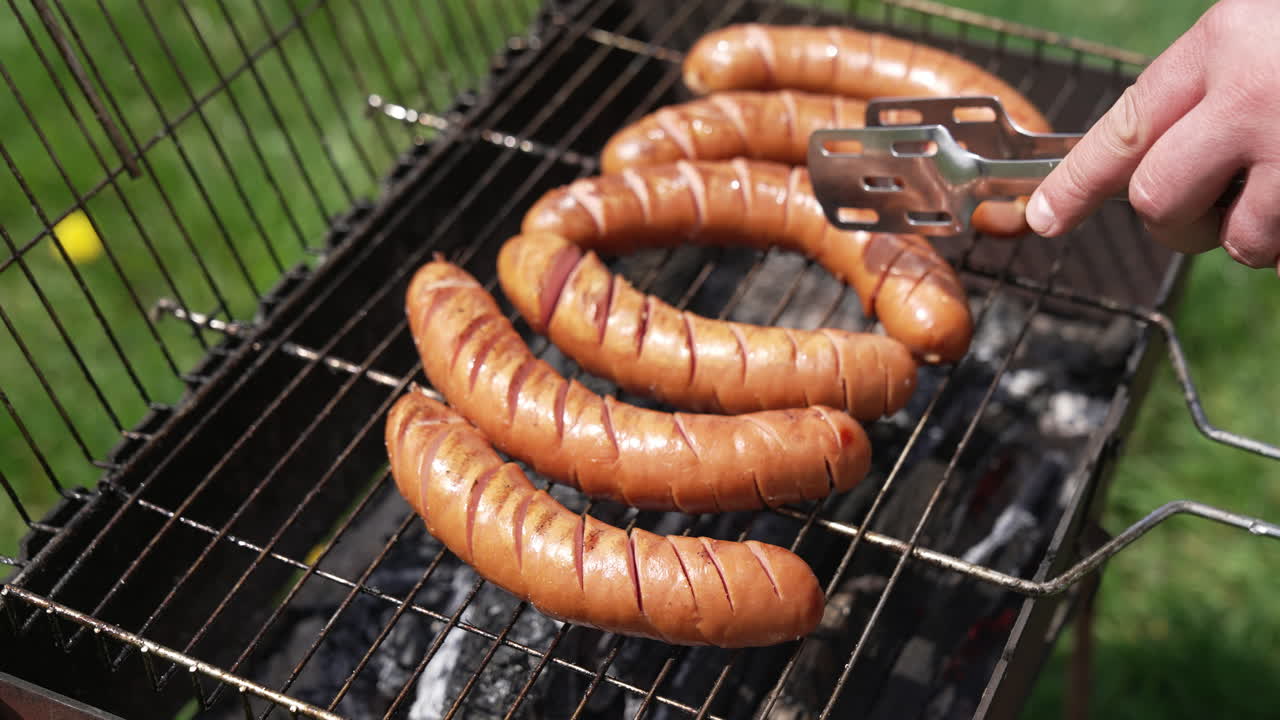 Tasty juicy sausages grilling. Barbecue grill cooking outdoors. Hot dog sausages are turning with tongs on a grill plate. Food for picnic.