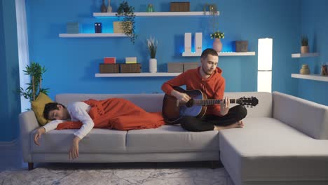 bored and sad young man at home playing guitar waking his younger brother from sleep.