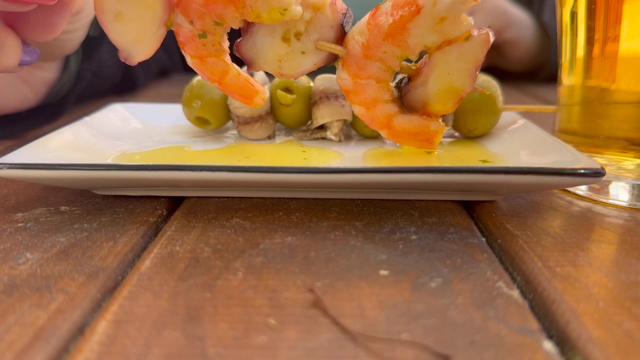 Vista ravvicinata delle mani di una giovane donna caucasica con le unghie dipinte di colori diversi che mangia tapas di pesce e beve birra in una terrazza.
