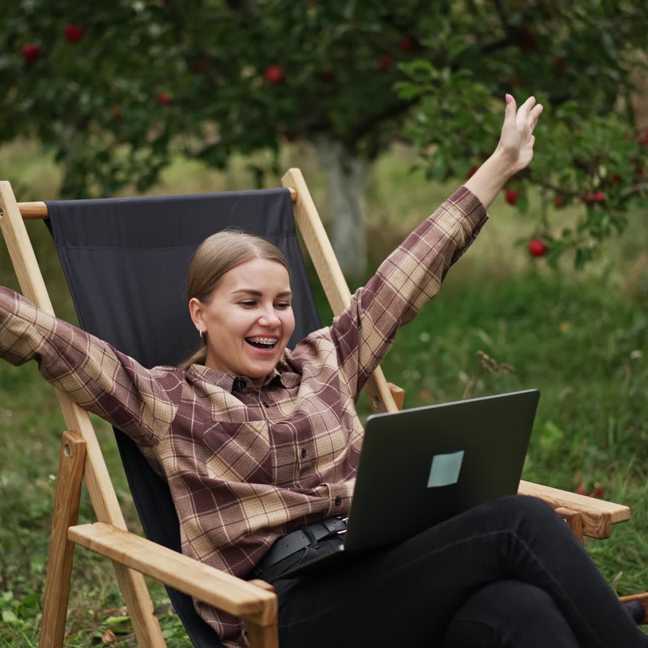 Cheerful lady having online video chat and is happy about something. Woman rejoices raising her hands and smiling happily. Nature at backdrop