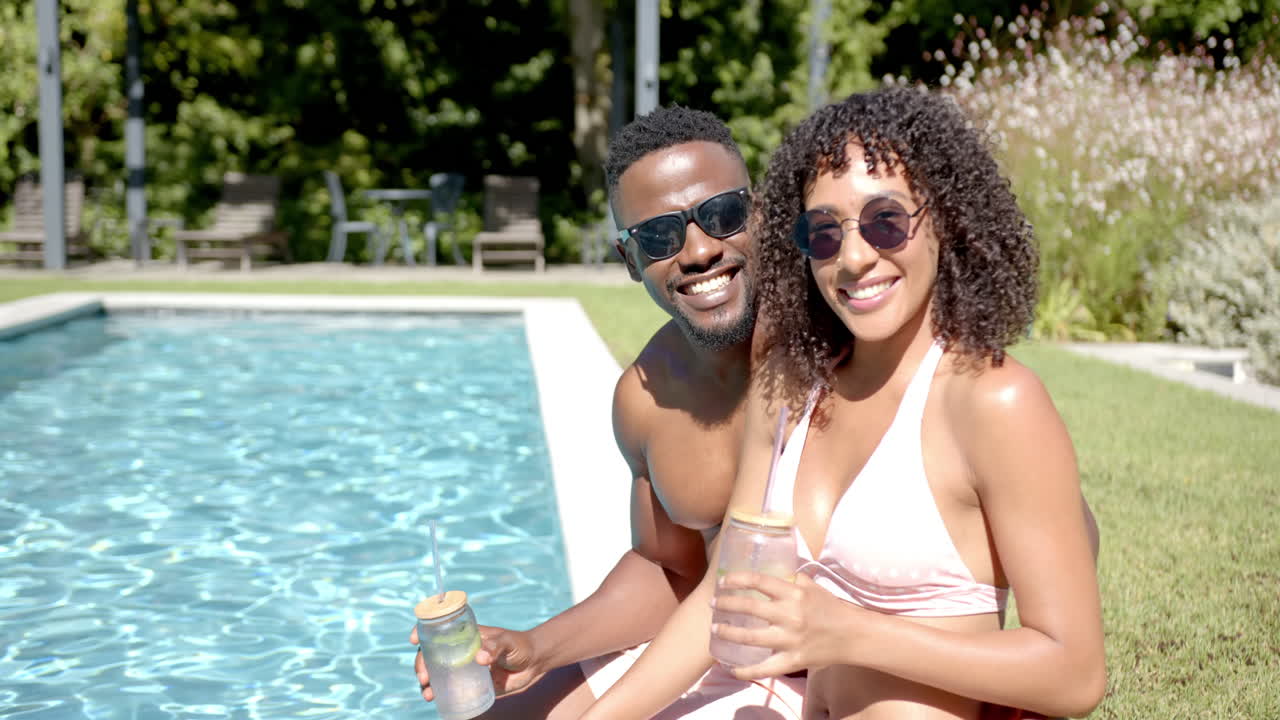 Lächelndes afroamerikanisches Paar entspannt sich am heimischen Pool, hält Getränke und genießt den sonnigen Tag.