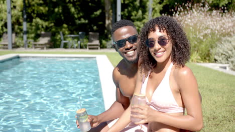 smiling african american couple relaxing by home pool, holding drinks and enjoying sunny day