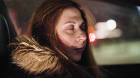 first-person view of a woman in a winter coat with a fur hood, inside a car at night. she has removed her head warmer, revealing her hair. the background shows blurry city lights