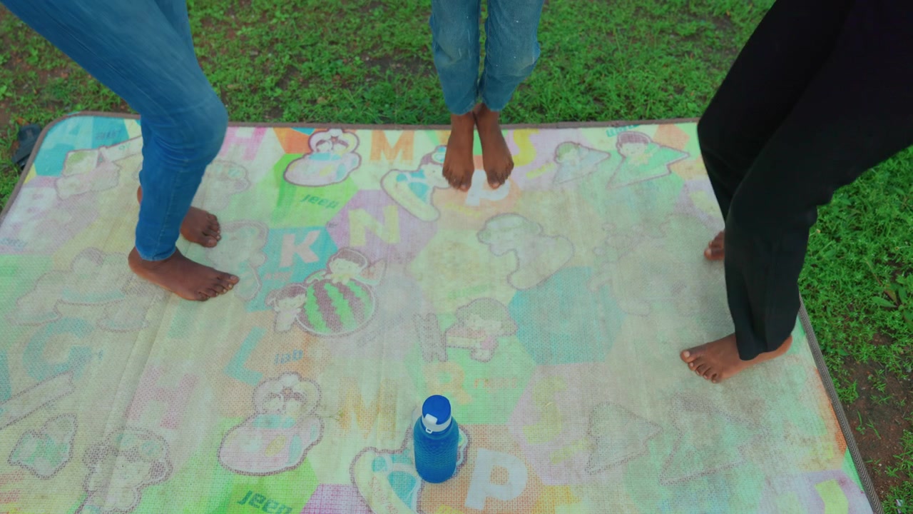 Mère et enfant pieds nus sur un tapis de craie, gros plan des jambes et des pieds marchant sur des dessins colorés sur le tapis, jean et leggings noirs, bouteille d'eau sur le bord, pelouse ensoleillée du parc, moment de complicité ludique.