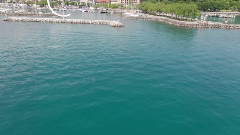 Drone-Volando-Sobre-El-Hermoso-Lago-Verde-Azulado,-Inclinándose-Hacia-Arriba-Y-Revelando-Un-Puerto-De-Yates-En-Lausana,-Suiza