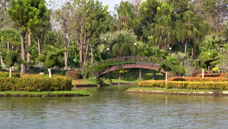 uma fonte pulverizando água em uma lagoa com uma ponte em arco ao fundo cercada por belas plantas