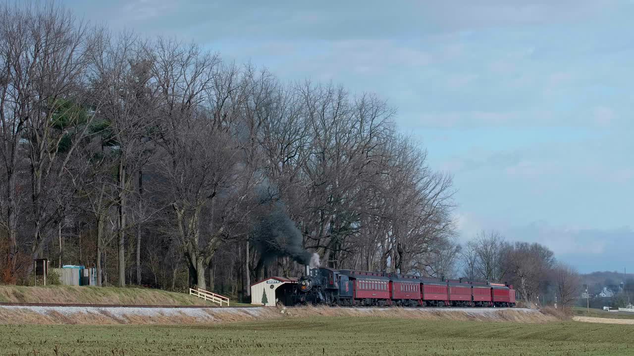 A steam train passes by a quiet rural station surrounded by trees and open fields. The peaceful landscape showcases the charm of the countryside in early spring.