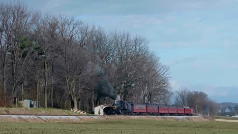 a steam train passes by a quiet rural station surrounded by trees and open fields. the peaceful landscape showcases the charm of the countryside in early spring.