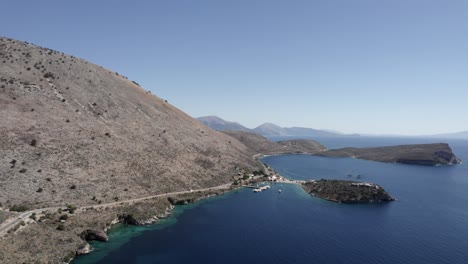 Drone-video-over-the-turquoise-waters-of-the-Albanian-coast-in-Porto-Palermo,-from-the-front-plane-advancing-over-the-island-of-Porto-Palermo-and-the-marina-with-yachts-and-boats