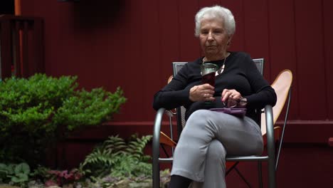 anciana sonriendo y sentada afuera en un jardín mientras bebe vino tinto