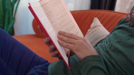 nahaufnahme einer person in grünem langarmhemd und blauen jeans, die auf einer orangefarbenen couch sitzt und ein rotes buch liest