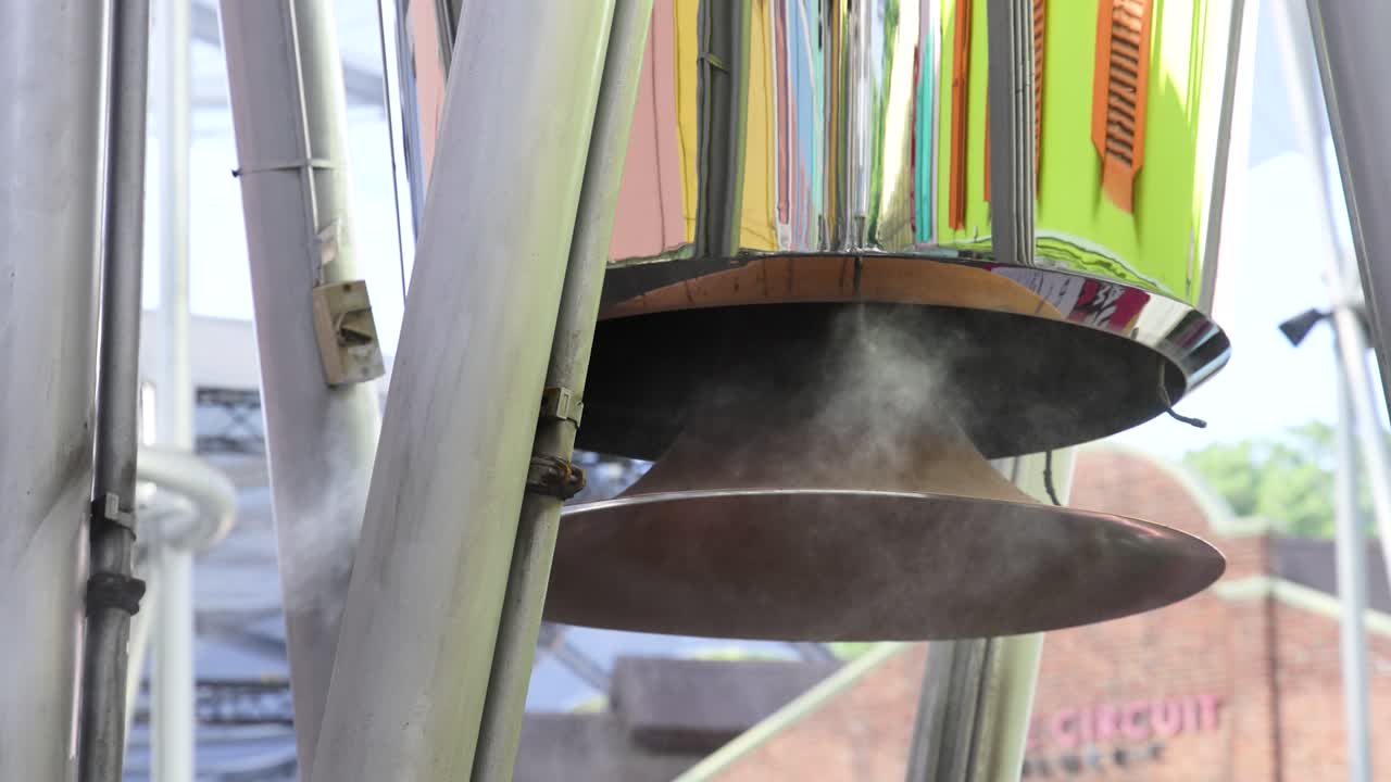 Close-up of a metallic exhaust vent releasing steam outdoors, surrounded by colorful urban architecture in daylight. Static camera, soft natural lighting