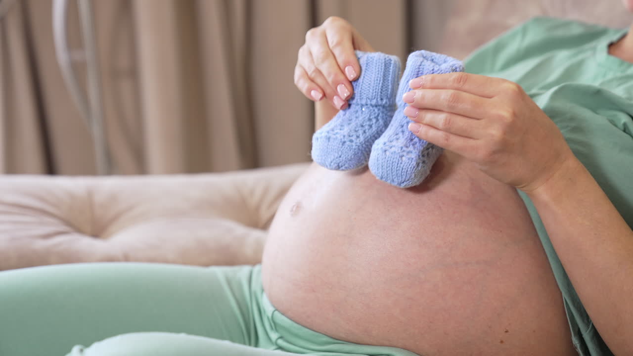 Tummy of a pregnant woman where she puts cute newborn socks and walks with them. Expecting mother playing with little baby socks on her tummy.