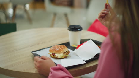 back view of woman in pink dress at a table, eating from a black tray containing a burger, fries, and coffee, she picks up a potato chip from the fries and enjoys her meal with a relaxed posture