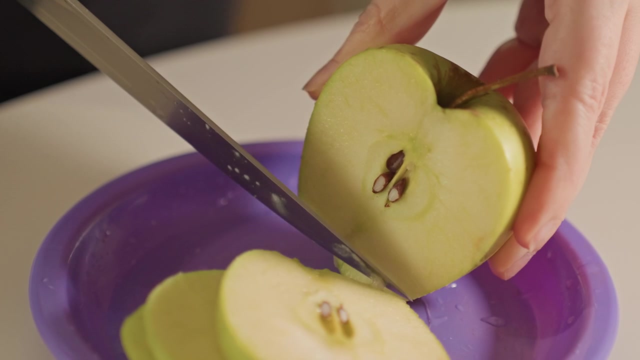 Primo piano di mani che affettano mele verdi a pezzi sottili. Le fette di mela cadono direttamente da dove sono state tagliate, accumulandosi. Piano di lavoro della cucina con superficie bianca visibile in alcune inquadrature