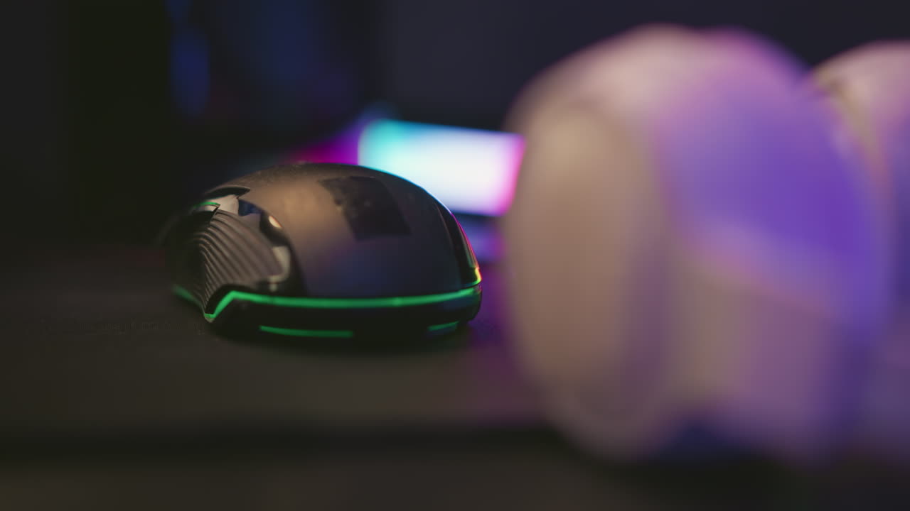 Close up black gaming mouse and white headset positioned on dark surface beside vibrant multicolor LED light bar creating dynamic contrast with ambient glow and tech-focused studio desk layout