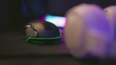close up black gaming mouse and white headset positioned on dark surface beside vibrant multicolor led light bar creating dynamic contrast with ambient glow and tech-focused studio desk layout