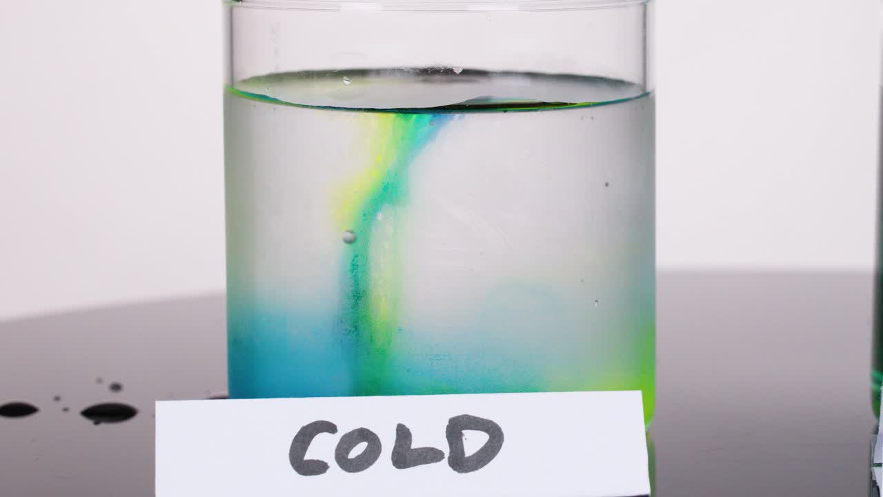 Colored dye slowly disperses in a clear glass of cold water under even studio lighting