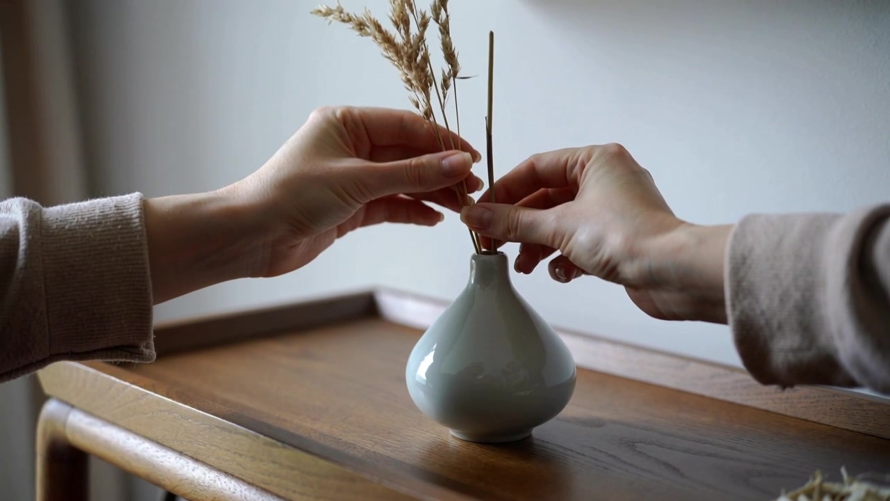 Entering frame hands arranging dried-stems into vase on tray by window for balance, wearing sweater