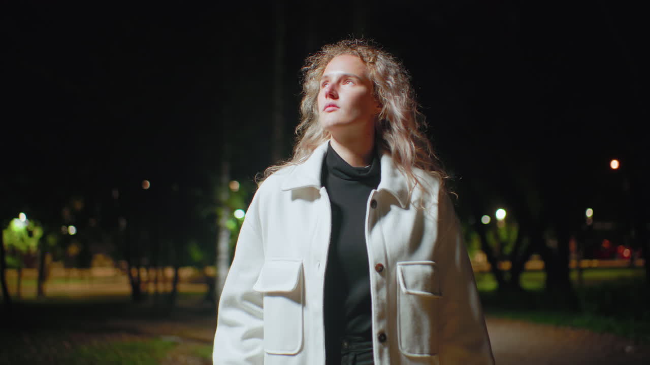 Young woman wearing white coat glances thoughtfully into background under night sky, surrounded by faint glowing park lights and trees, standing still with contemplative expression