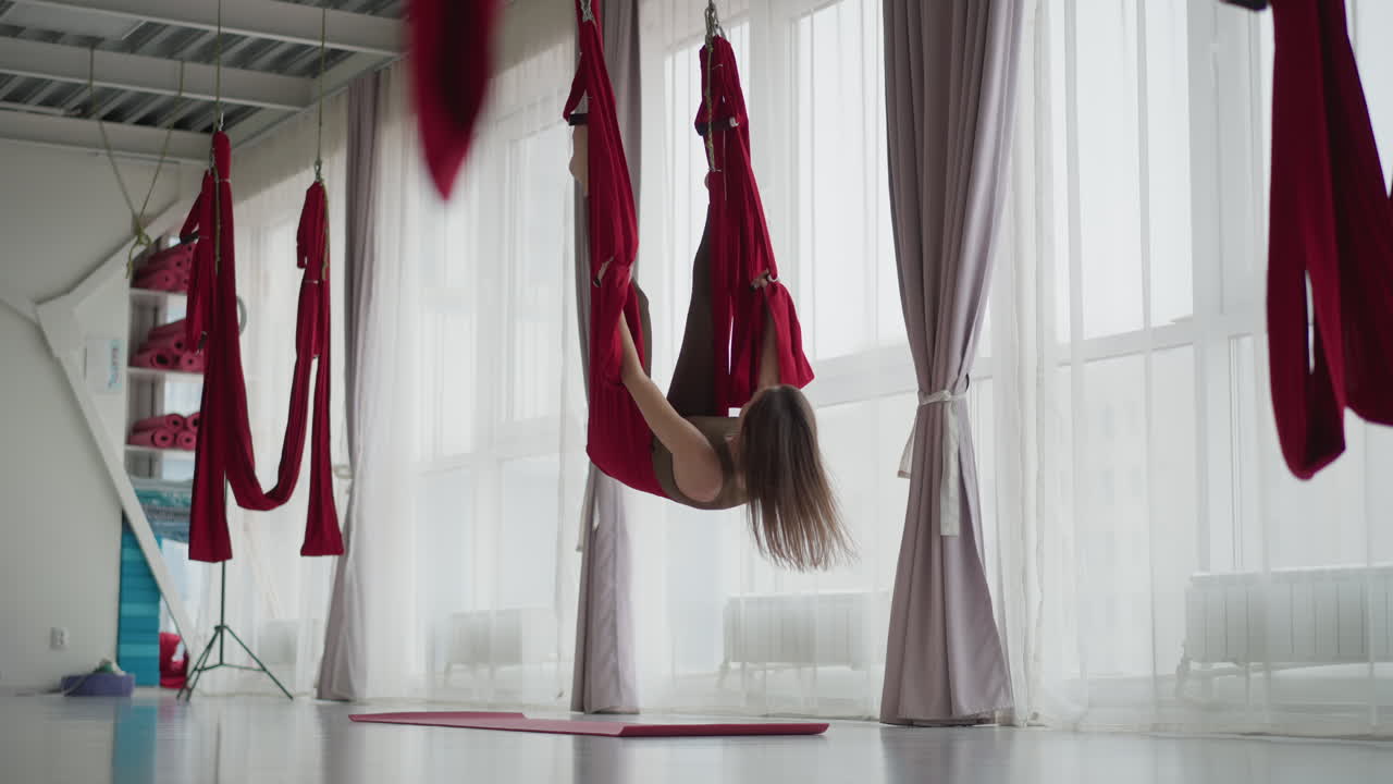 Wellness guide gently swings in red silk hammock near floor to ceiling windows, legs suspended mid air while adjusting grip on fabric handles, bright airy studio environment calming graceful movement