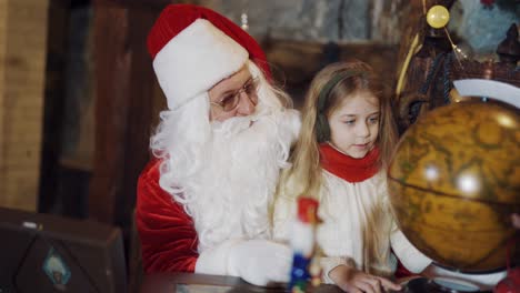 santa claus and beautiful girl look at the round globe indoors. child's hands turning the globe model and santa explaining to a little girl something.