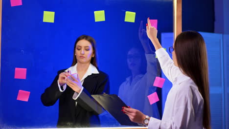 long-haired brunette ladies stand near the glass wall. woman writes on the stickers and puts them on the board. planning concept.