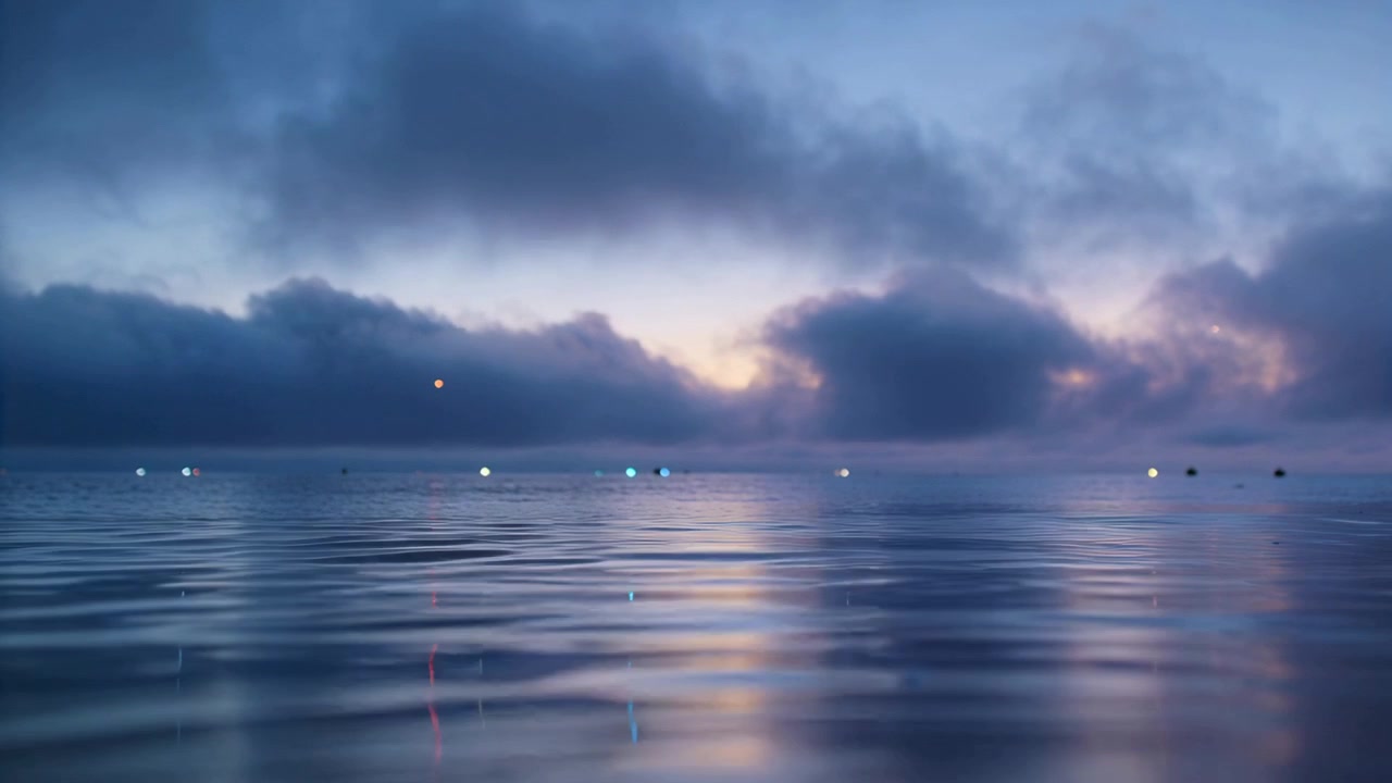 Rippling calm water surface reflecting sunrise glow as clouds drift at sea, with horizon lights